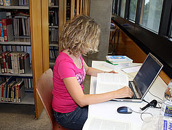 Das Bild zeigt einen Arbeitsplatz in der Uni-Bibliothek. Eine bwin_bwinֹӭ@ sitzt an einem Tisch und liest ein Buch. Auf dem Tisch steht ein Laptop. Man sieht auf dem Tisch auch eine Flasche Wasser und mehrere Bcher.